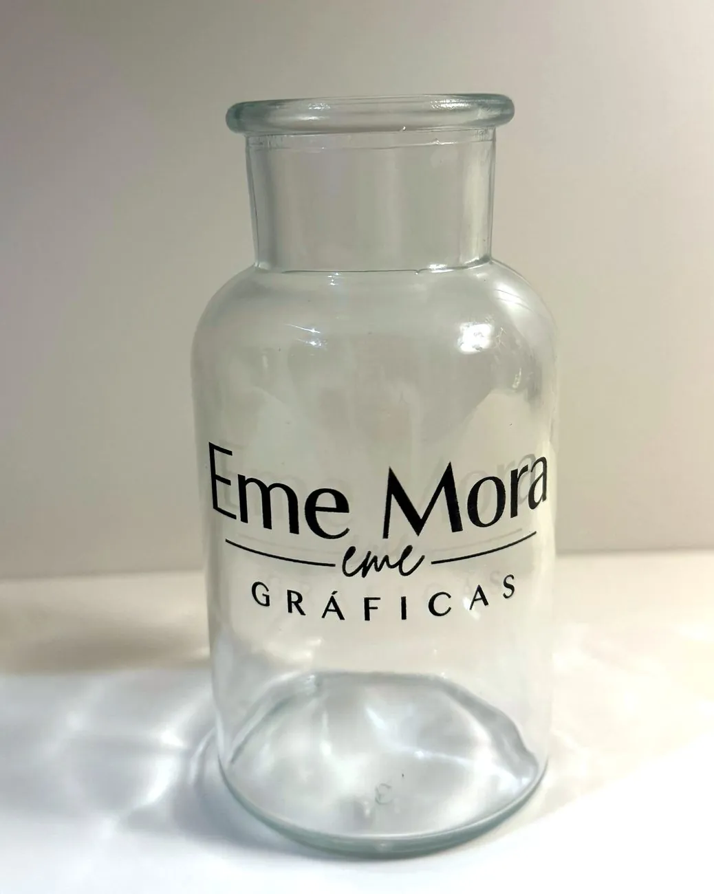 Frasco de vidrio transparente con el logo de Gráficas Eme Mora impreso en negro